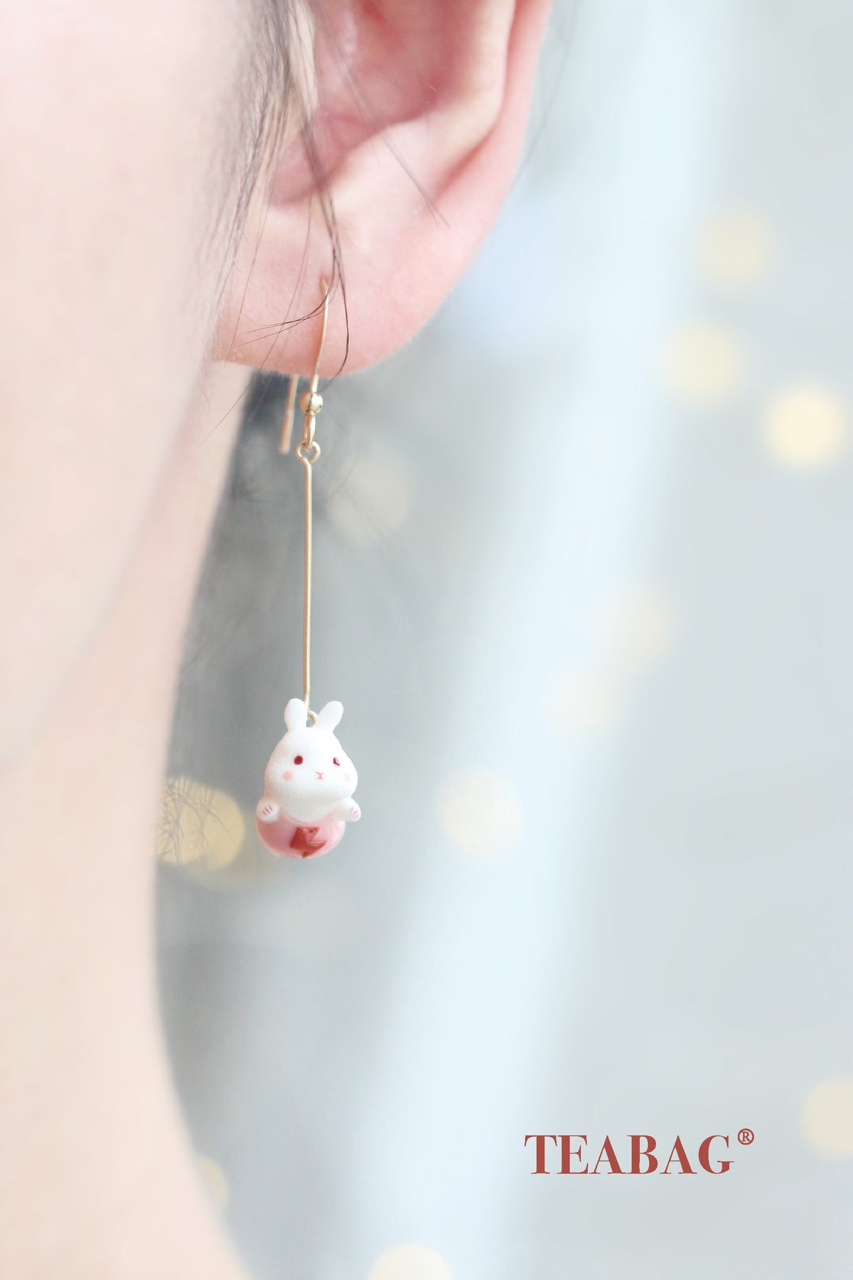 Flying Bunny long pierced-hook earring worn on an ear, showing the complete straight extension, pink outfit and golden cape with TEABAG branding. Sold as a pair.