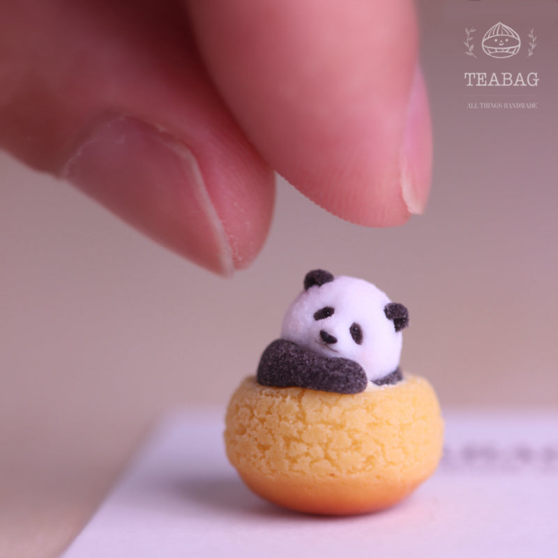 Complete front view of the Resting Panda Cream Puff miniature without jewelry fittings, showing the resting panda, full golden pastry, fingertip scale reference and TEABAG logo in a square photograph.
