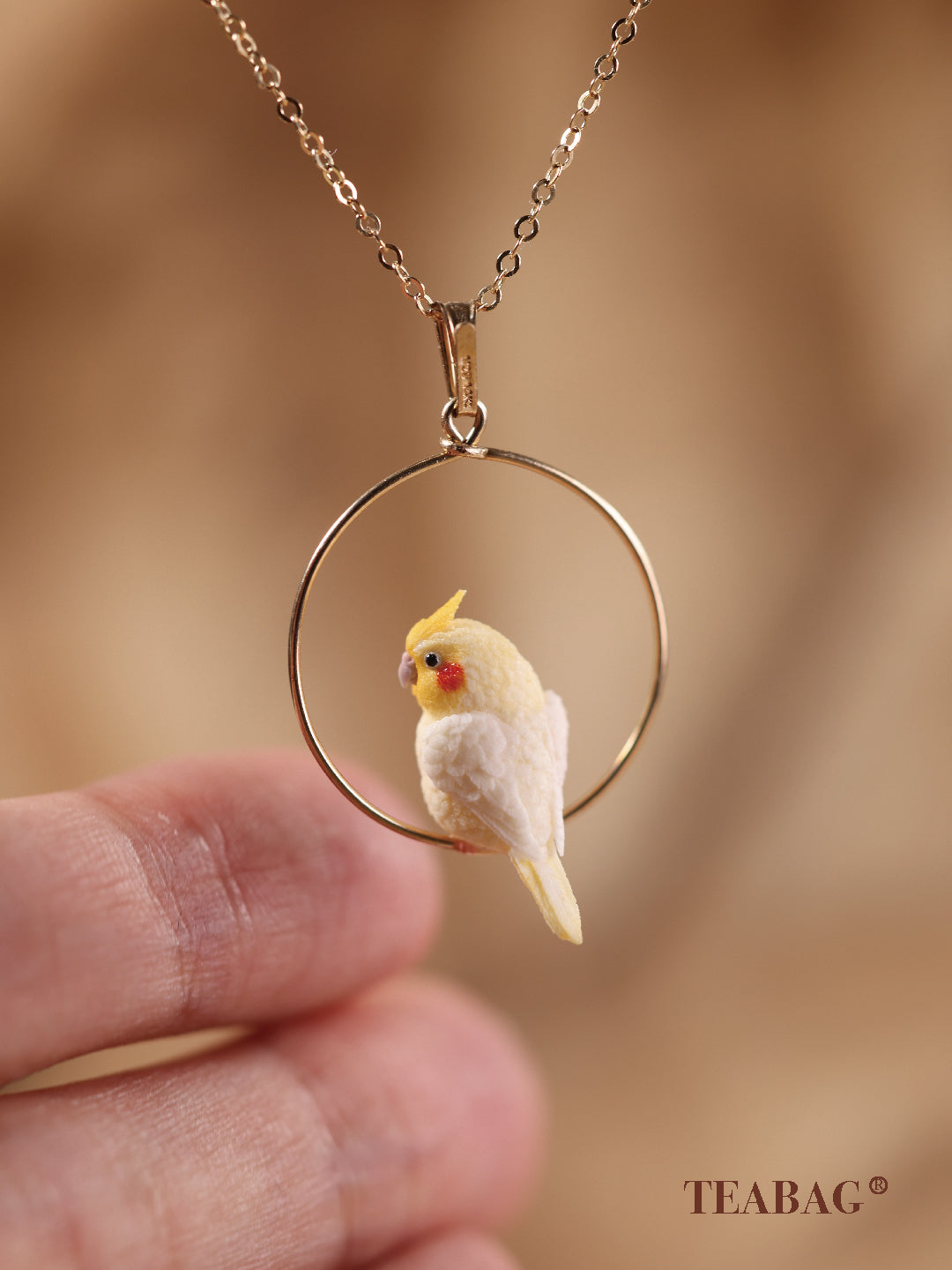 Side and back view of the cockatiel necklace pendant, showing the layered wings and tail, complete hoop and bail, with a fingertip for scale.