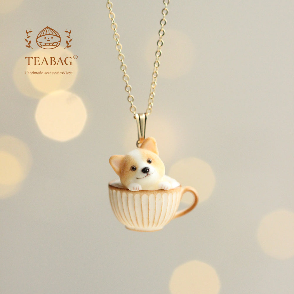 Corgi Coffee necklace pendant, with a tiny corgi peeking out of a ribbed cream-and-gold cup, showing the complete bail and chain against a warm background with TEABAG branding.