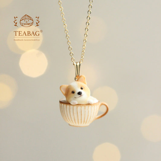 Corgi Coffee necklace pendant, with a tiny corgi peeking out of a ribbed cream-and-gold cup, showing the complete bail and chain against a warm background with TEABAG branding.