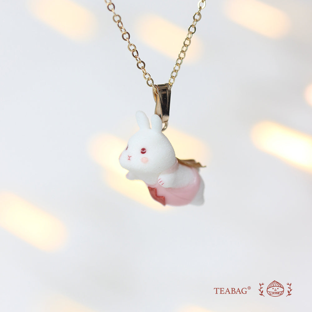 White flying-bunny pendant in pink with a gold-colored cape, bail and chain, shown in three-quarter view with TEABAG branding.
