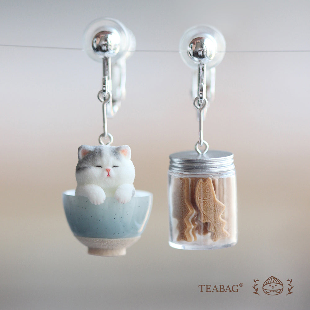 Complete Kitten Bakery clip-on earring pair: a gray-and-white kitten in a blue bowl and a miniature cookie jar, with both clip fittings and TEABAG branding visible.
