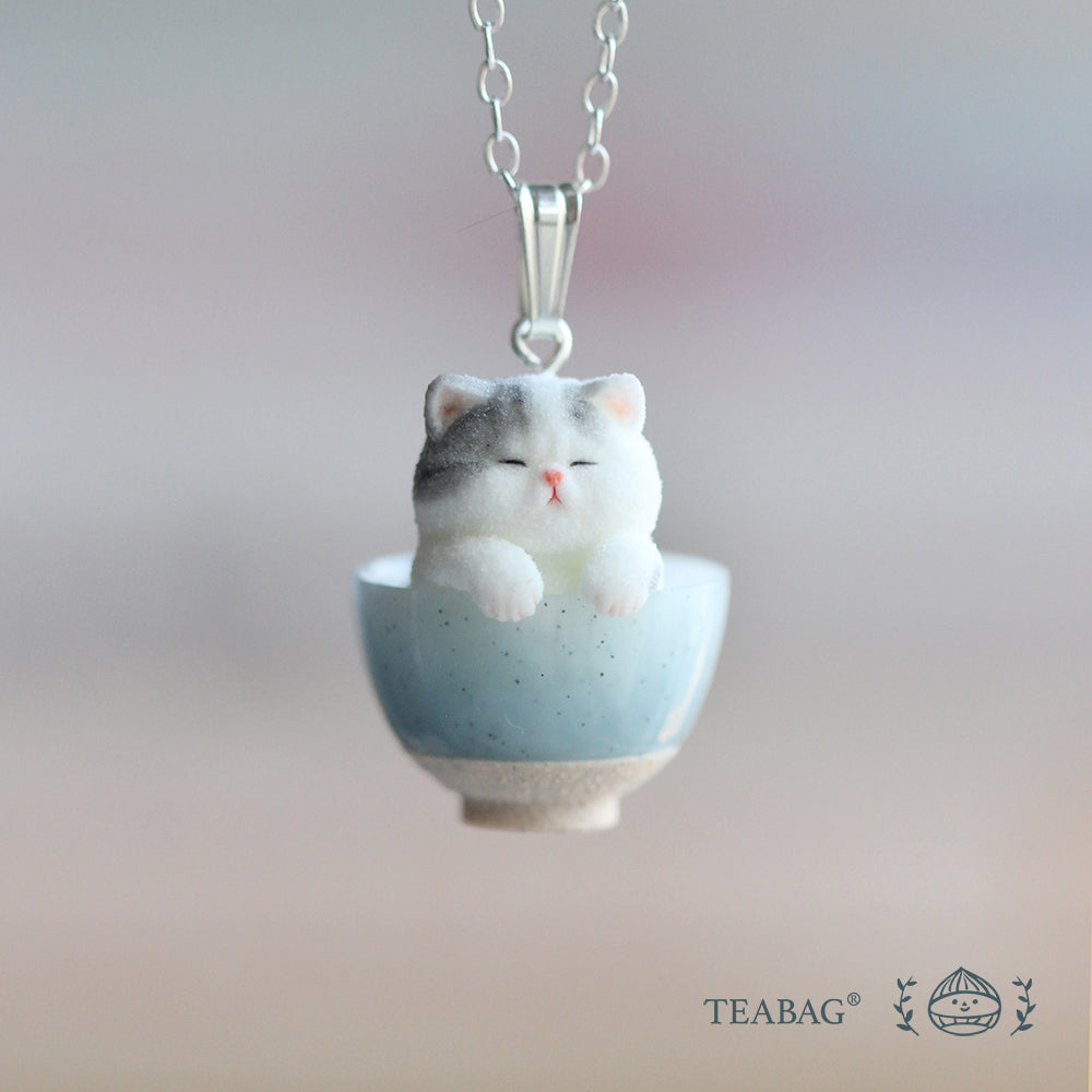 Front view of the Kitten Bakery necklace: a grey-and-white kitten in a blue bowl, with its bail and a section of chain visible. TEABAG logo.
