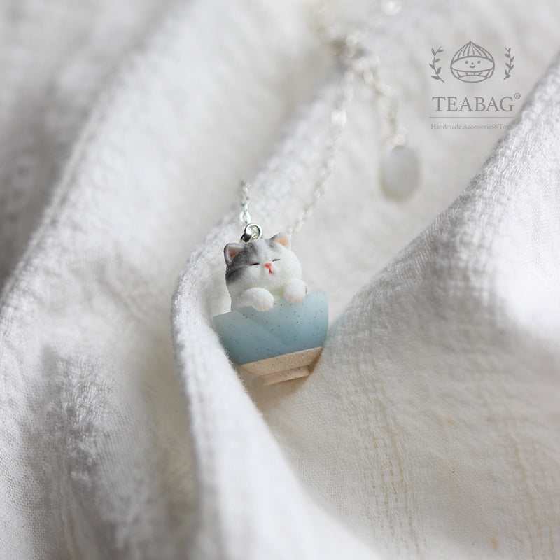 Kitten Bakery necklace resting on pale linen, showing the gray-and-white kitten in its blue bowl, bail and chain, with TEABAG branding.