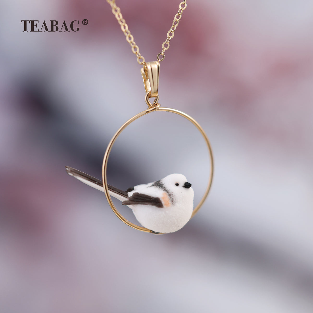 Side view of the white little-bird pendant with dark wings and a long tail inside its complete circular hoop, with bail, chain and TEABAG branding.