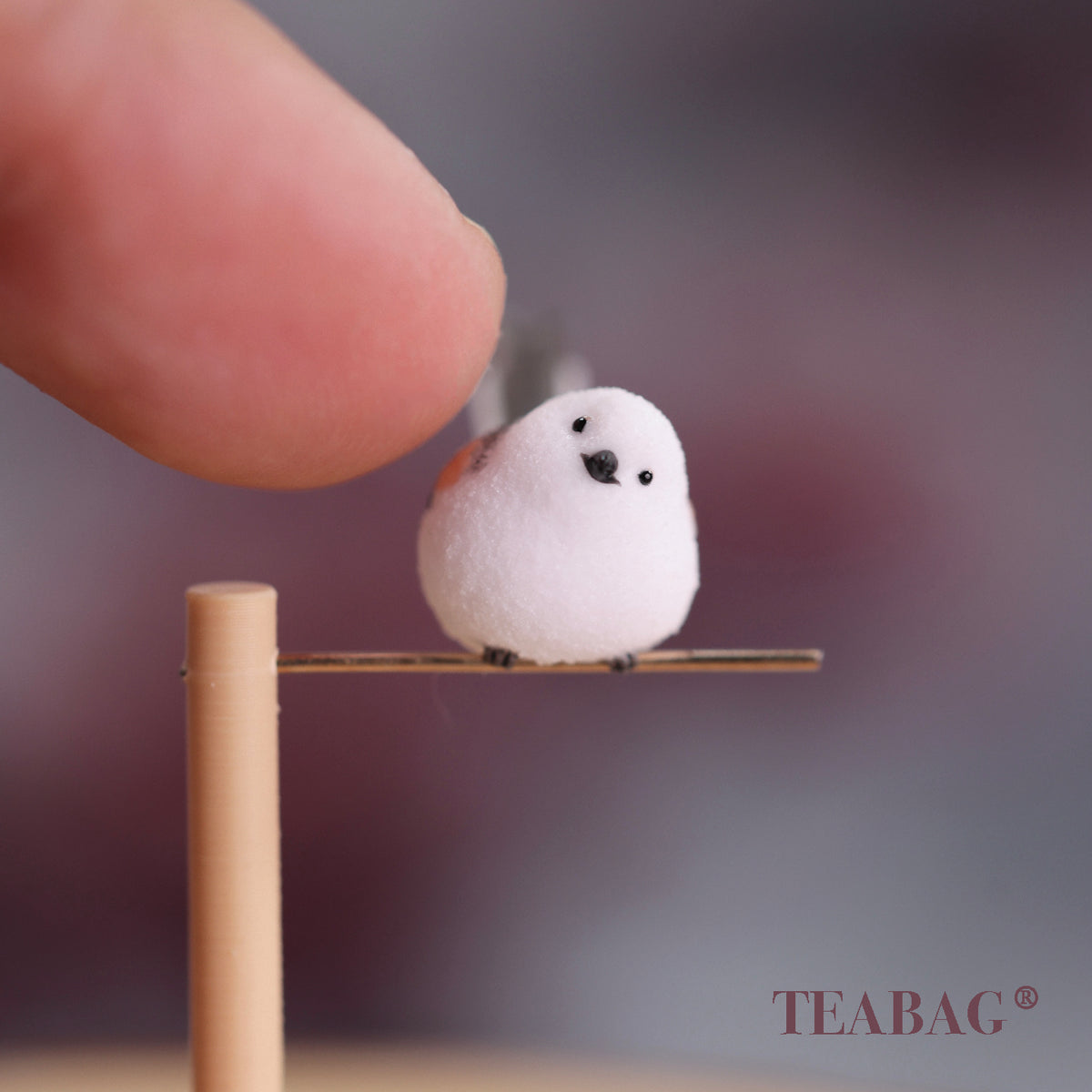 Tilted-head white bird miniature on a pine perch and gold-colored crossbar, with a fingertip for scale and TEABAG branding.