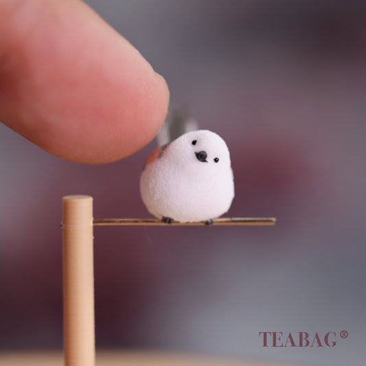 Tilted-head white bird miniature on a pine perch and gold-colored crossbar, with a fingertip for scale and TEABAG branding.