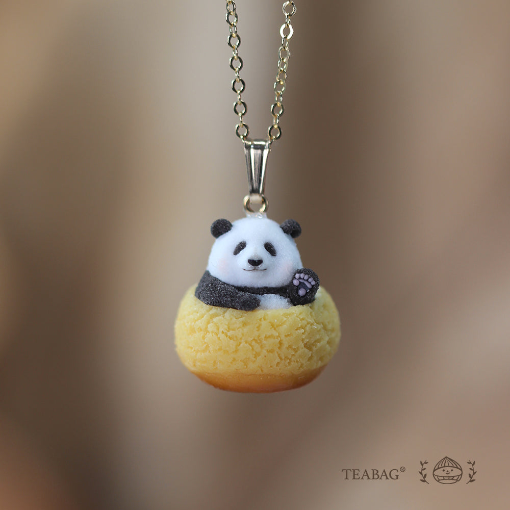 Waving panda cream puff pendant on a necklace chain, shown from the front against a warm brown background with TEABAG branding.