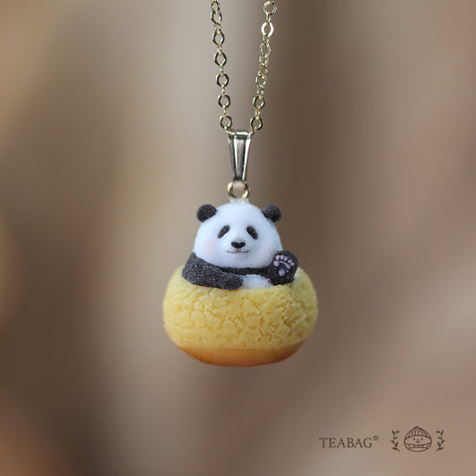Waving panda cream puff pendant on a necklace chain, shown from the front against a warm brown background with TEABAG branding.