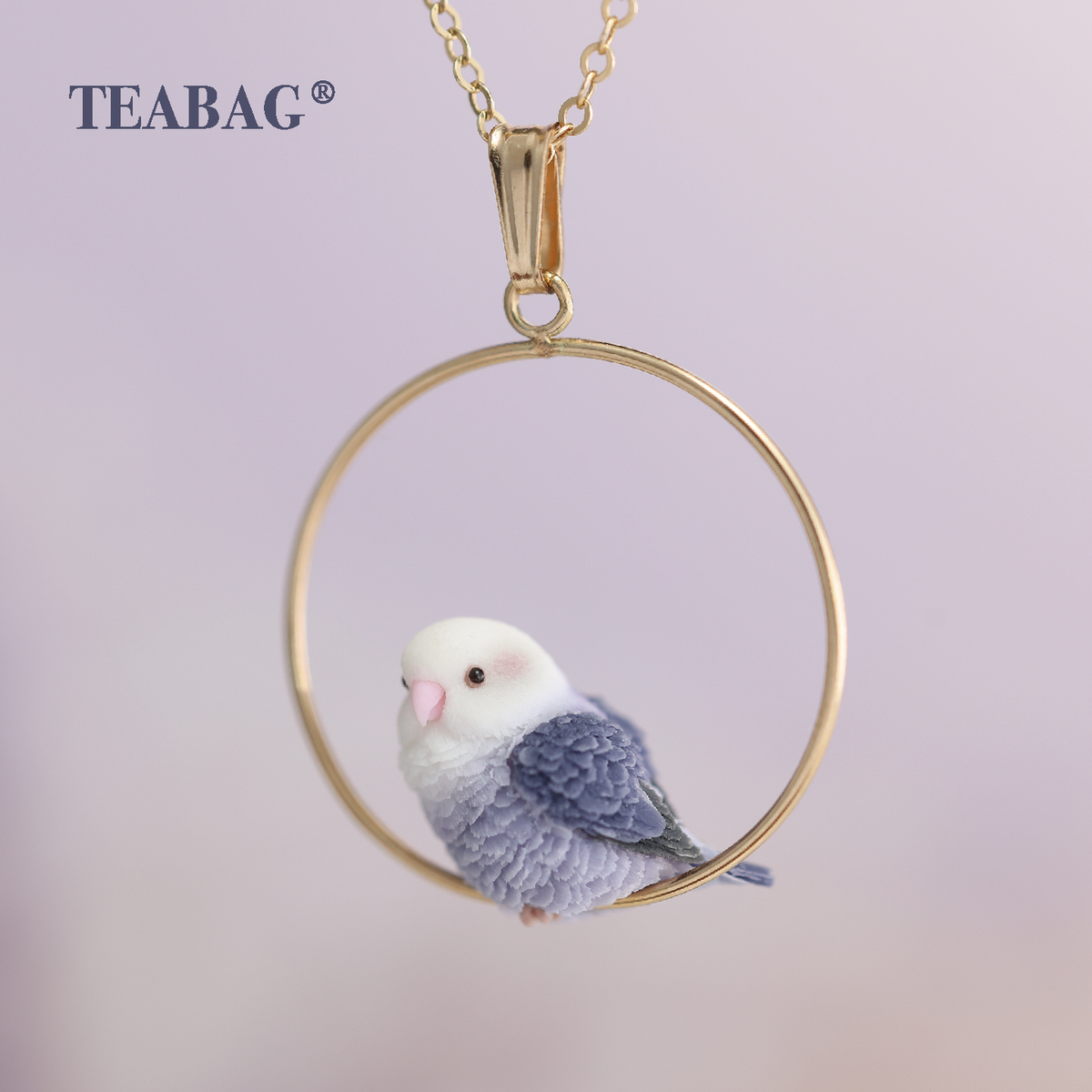Front view of the Violet Lovebird Necklace: a white-faced purple bird with a pink beak perched in a gold-colored hoop, with bail, chain section and TEABAG branding.