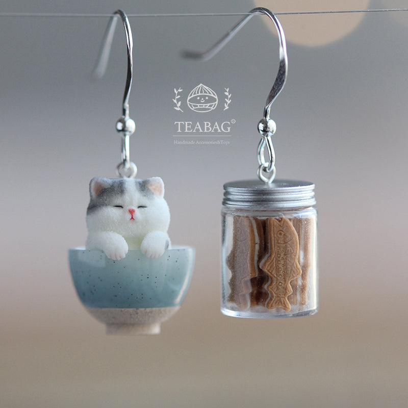 Complete Kitten Bakery pierced-hook earring pair: a gray-and-white kitten in a blue bowl and a miniature cookie jar, with both silver-colored hooks and TEABAG branding visible.