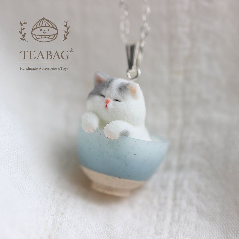Side detail of the Kitten Bakery necklace on pale linen, showing the kitten, blue bowl and silver-colored bail with TEABAG branding.