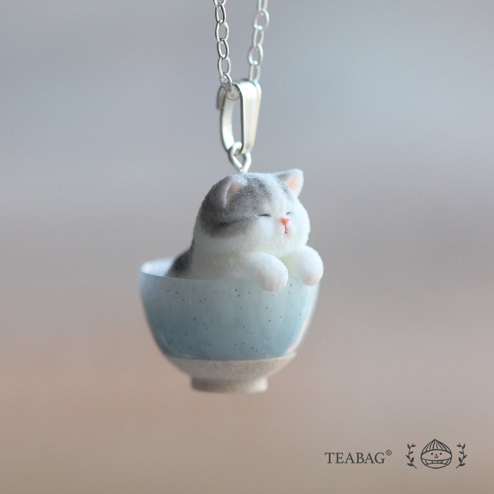 Three-quarter view of the Kitten Bakery necklace, showing the grey-and-white kitten in a blue bowl, its bail and a section of chain. TEABAG logo.