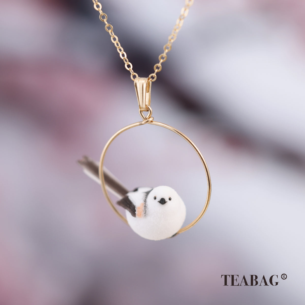 Front view of the white little-bird pendant with dark wings and a long tail inside its complete circular hoop, with bail, chain and TEABAG branding.