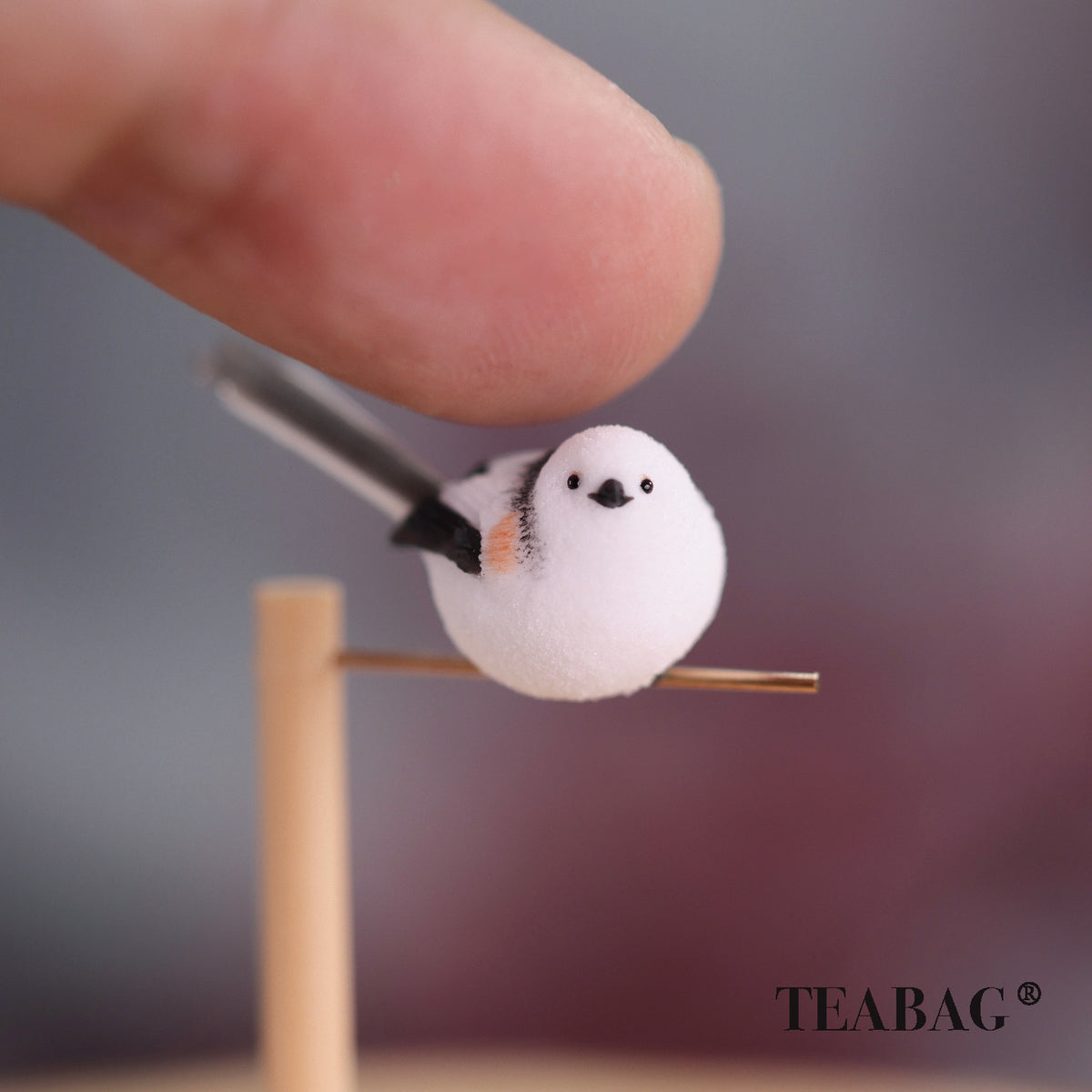 Side-facing white bird miniature on a pine perch and gold-colored crossbar, with a fingertip for scale and TEABAG branding.