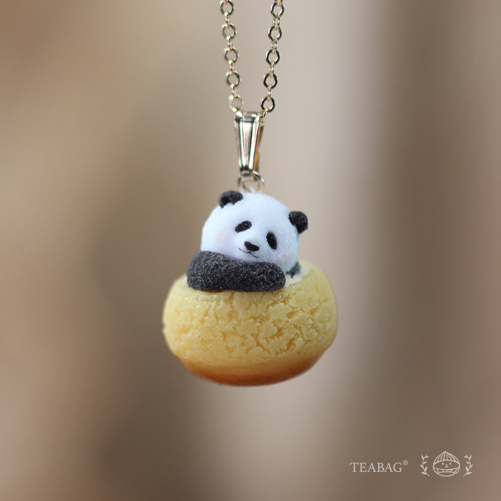 Resting panda cream puff pendant with a tilted head on a necklace chain, against a warm brown background with TEABAG branding.