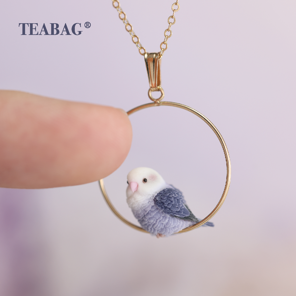 Violet Lovebird Necklace beside a fingertip for scale, showing the miniature purple bird, circular gold-colored hoop, bail and chain section, with TEABAG branding.