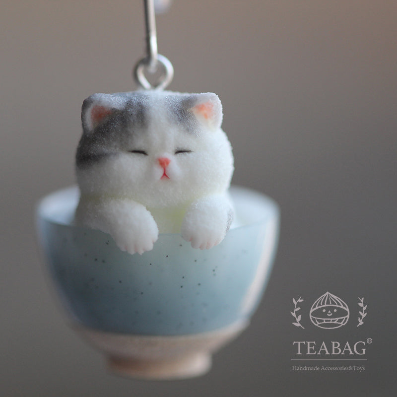 Close-up of the gray-and-white kitten earring charm resting its paws on a blue bowl, with TEABAG branding.
