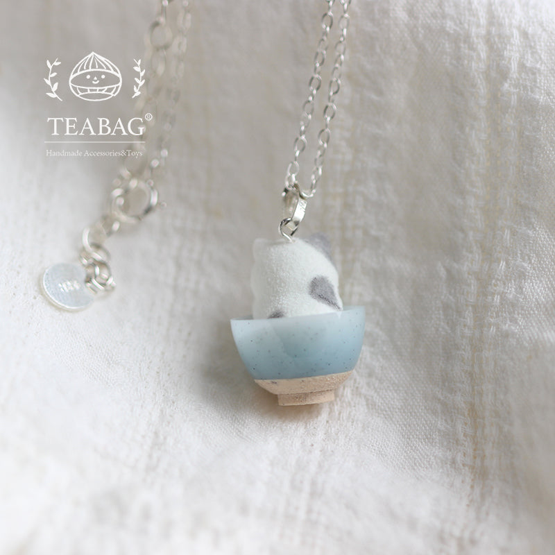 Rear view of the Kitten Bakery necklace on pale linen, showing the gray-and-white kitten in its blue bowl, bail and chain with TEABAG branding.