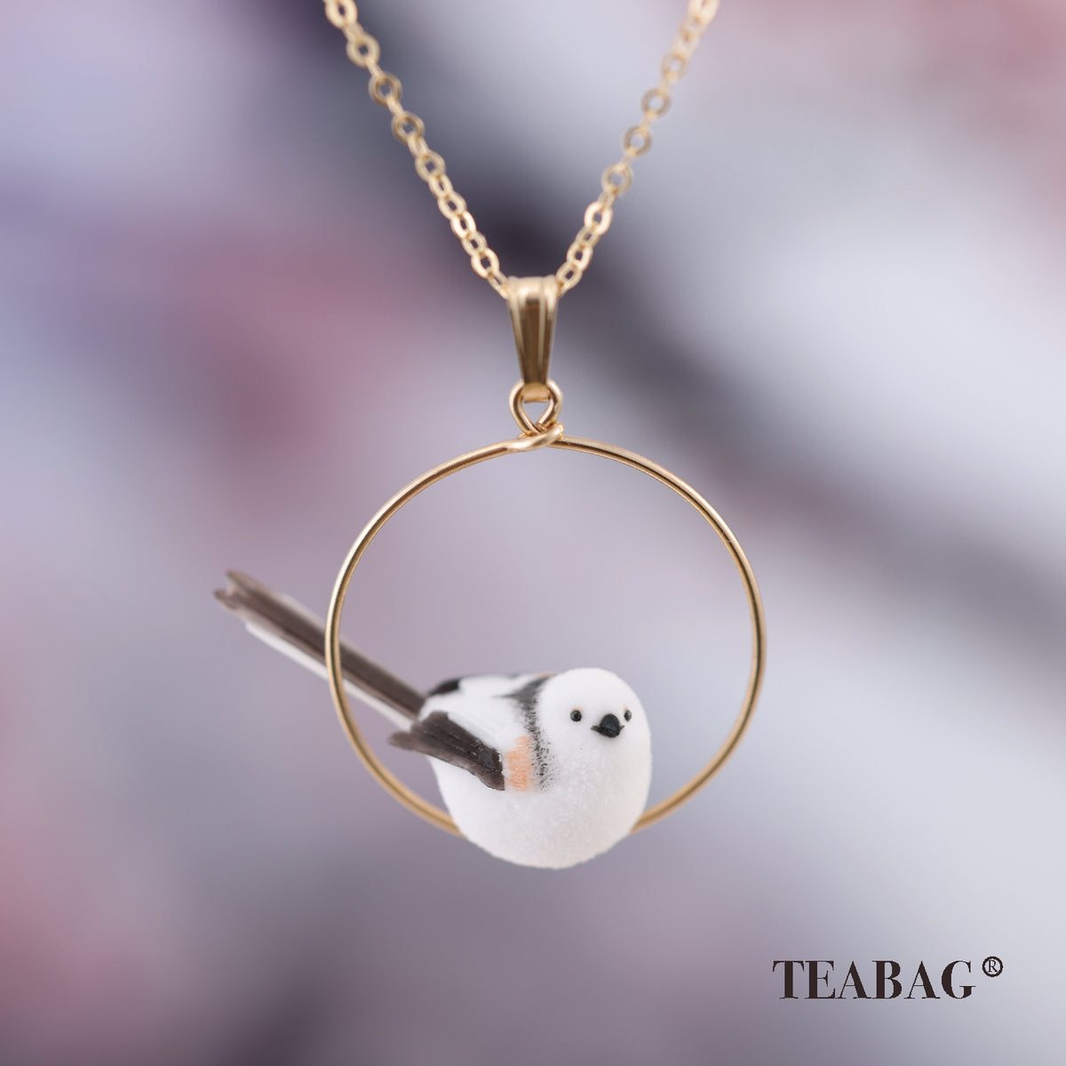 Three-quarter view of the white little-bird pendant with dark wings and a long tail inside its complete circular hoop, with bail, chain and TEABAG branding.