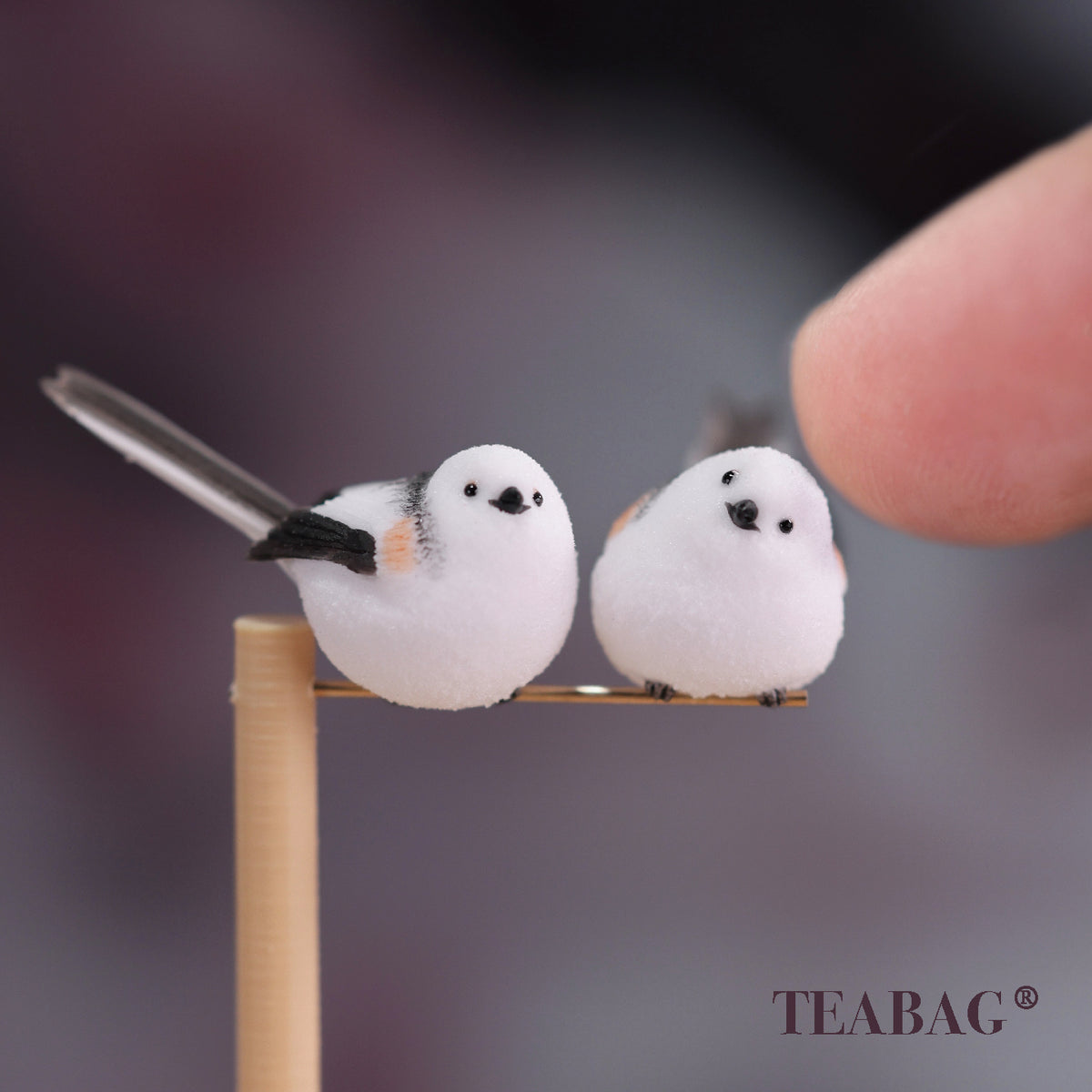 Pair of white bird miniatures, one side-facing and one tilted-head, sharing a pine perch and metal crossbar, with a fingertip for scale.