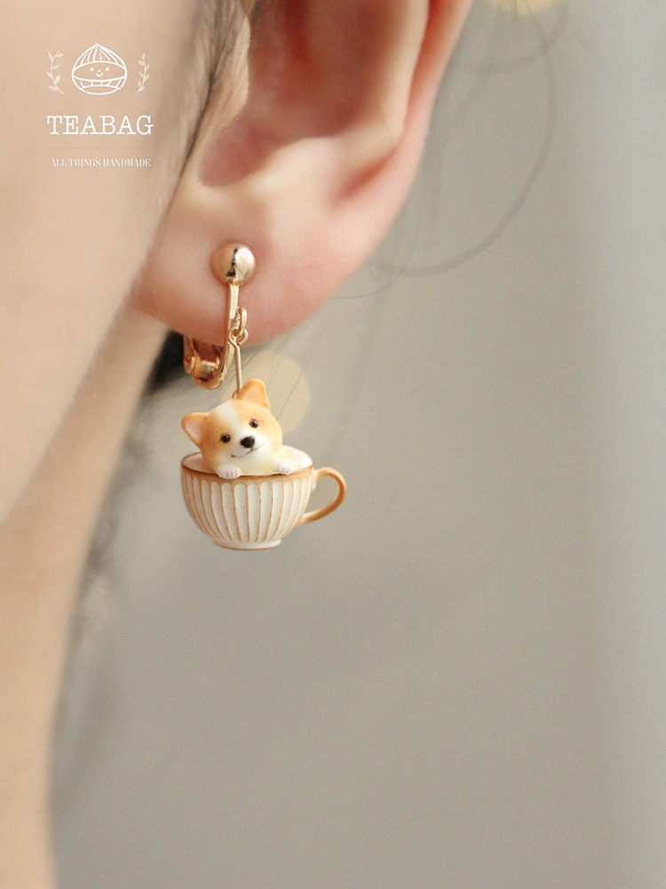 Corgi Coffee clip-on earring worn on an ear, showing the front-facing corgi in a ribbed cream-and-gold cup, the complete clip fitting and TEABAG branding.