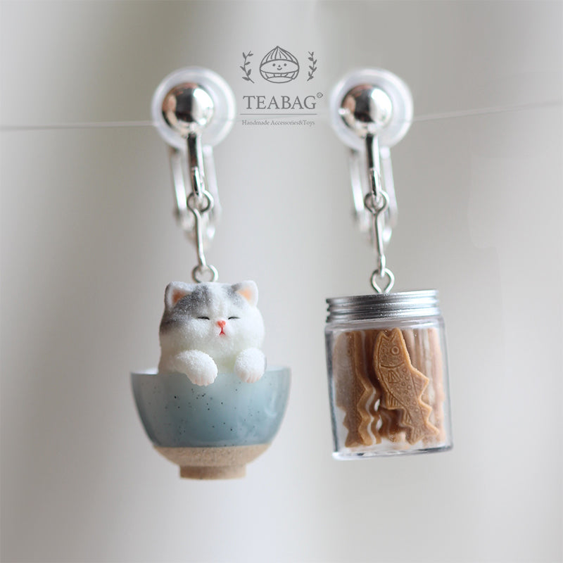 Front view of the complete Kitten Bakery clip-on earring pair, showing the gray-and-white kitten in a blue bowl, miniature cookie jar, both silver-colored clip fittings and TEABAG branding.