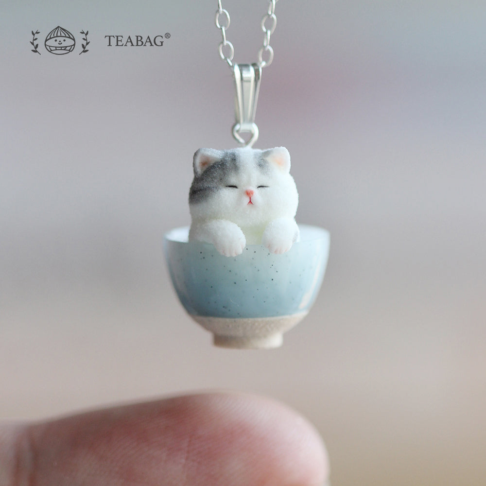 Kitten Bakery necklace pendant suspended above a fingertip, with the kitten in a blue bowl, its bail and a section of chain visible. TEABAG logo.