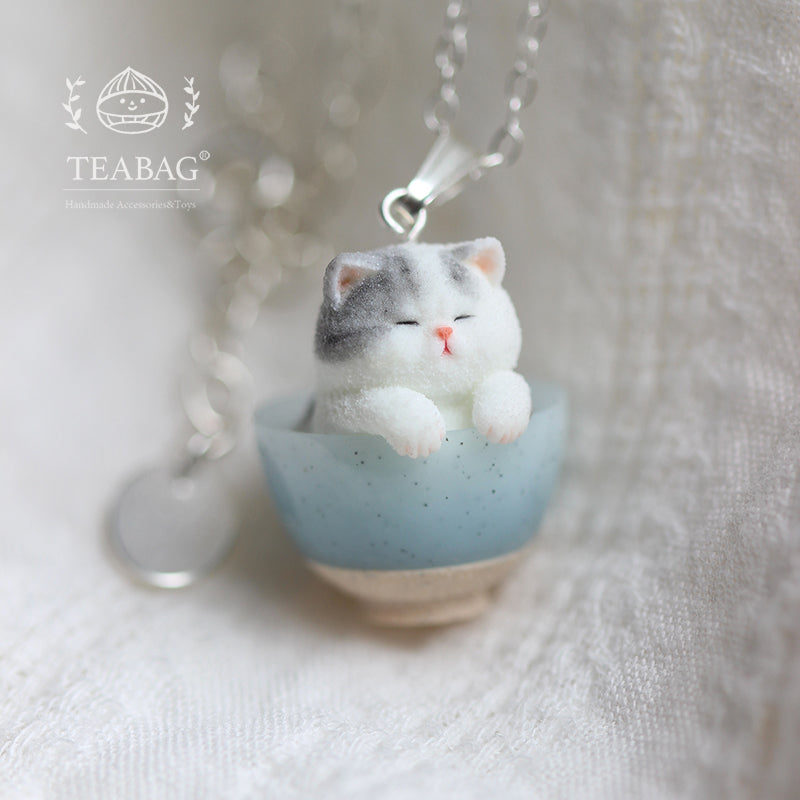 Front close-up of the Kitten Bakery necklace on pale linen, showing the gray-and-white kitten resting its paws on a blue bowl, with bail, chain and TEABAG branding.