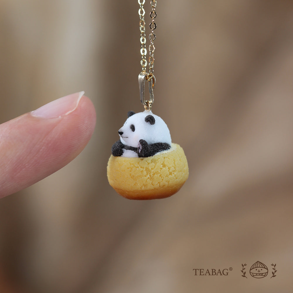 Side view of the waving panda cream puff necklace pendant beside a real fingertip for scale, with TEABAG branding.