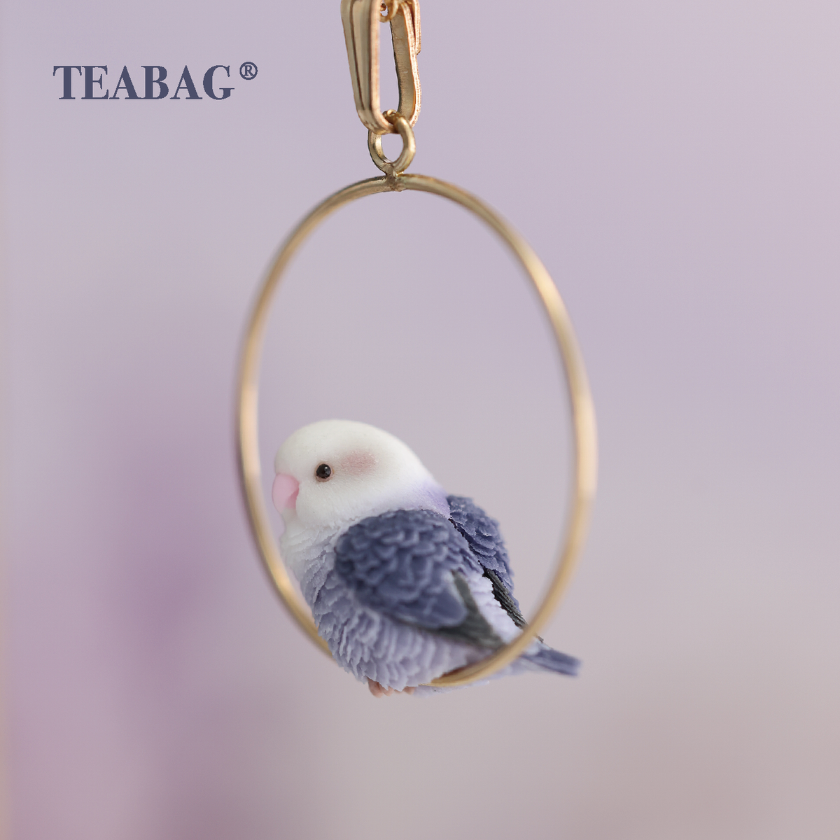 Side and back detail of the Violet Lovebird Necklace, showing the pale face, layered purple wings and tail within the gold-colored hoop, with TEABAG branding.