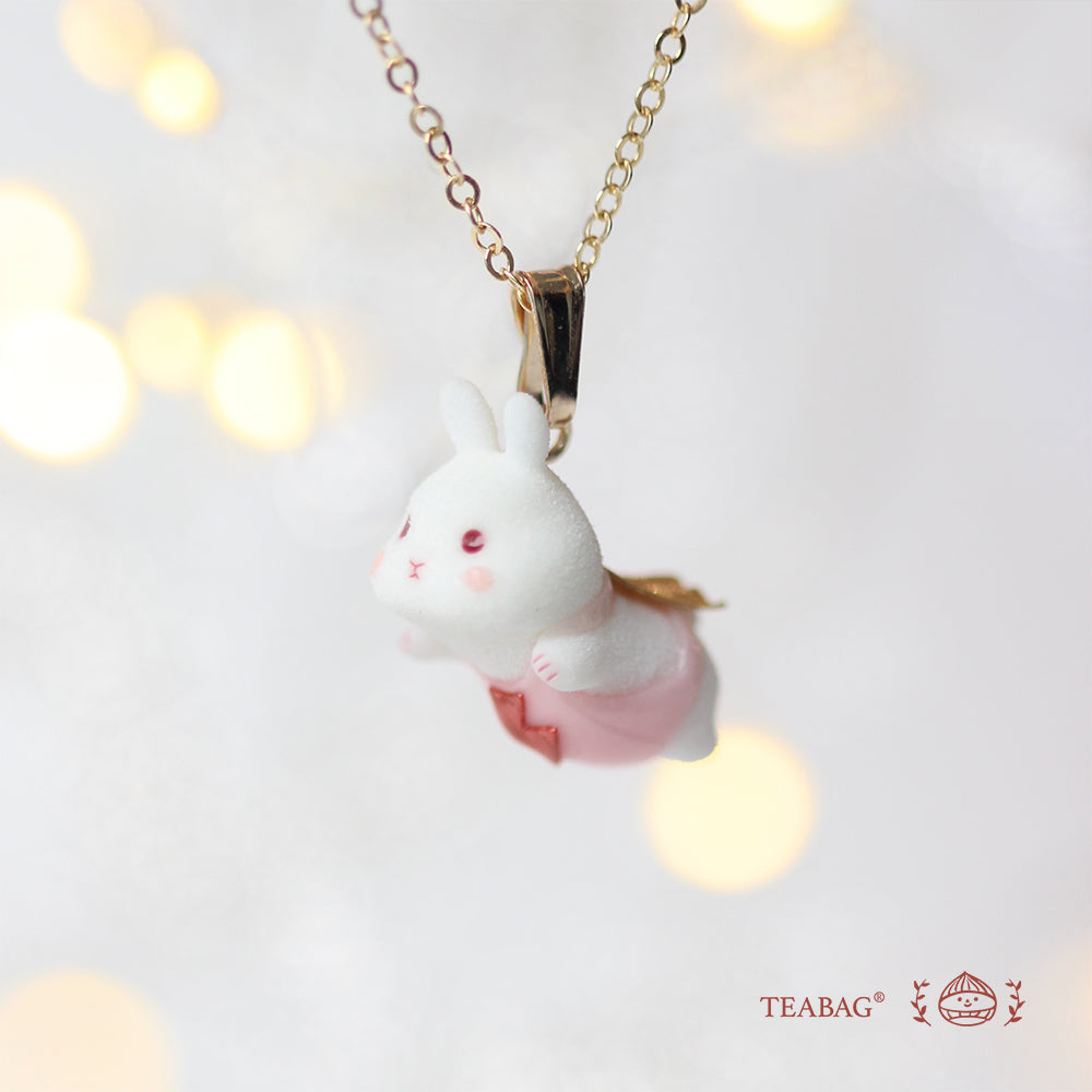 Side view of the white flying-bunny pendant in pink with its cape, bail and chain against warm light spots, with TEABAG branding.