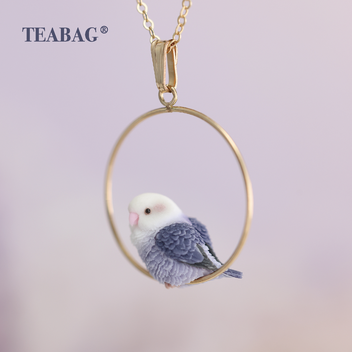 Side view of the Violet Lovebird Necklace, showing the white head, pink beak and sculpted purple feathers, with the full gold-colored hoop, bail, chain section and TEABAG logo.