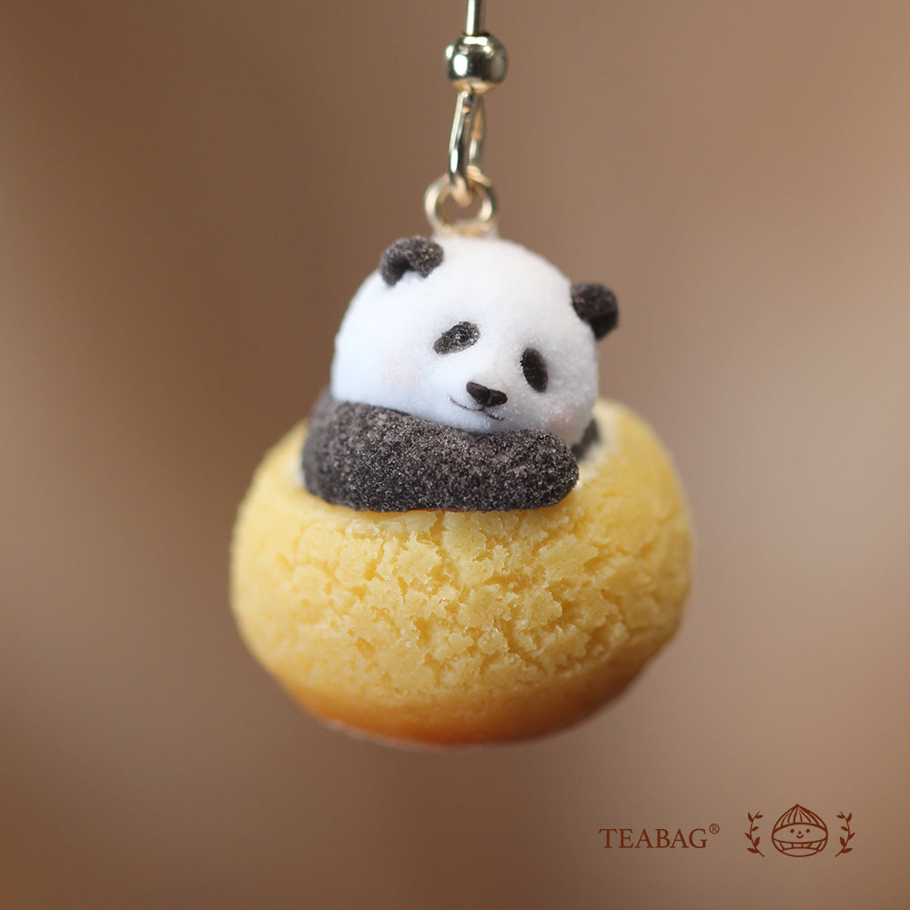 Close-up of a resting panda nestled in a textured golden cream puff, showing its tilted head and earring connector against a warm brown background with TEABAG branding.