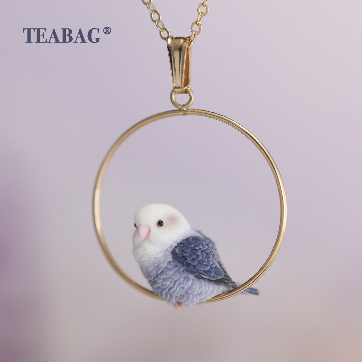 Angled front view of the Violet Lovebird Necklace, showing the pale face, pink beak and lavender feathered body inside the complete gold-colored hoop, with bail, chain and TEABAG branding.