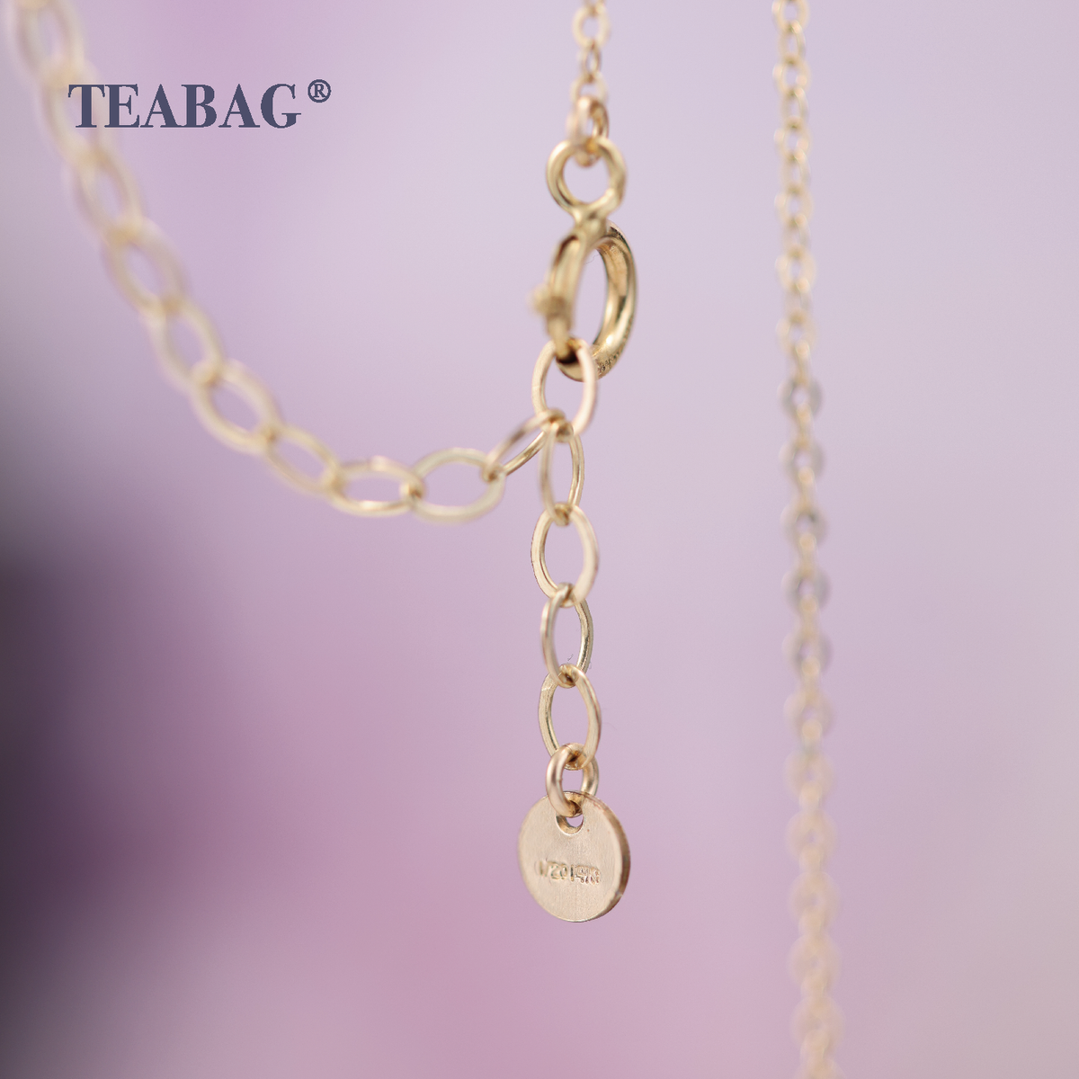 Detail of the Violet Lovebird Necklace chain, showing the gold-colored spring-ring clasp, extension links and small round end tag against a lavender background, with TEABAG branding.
