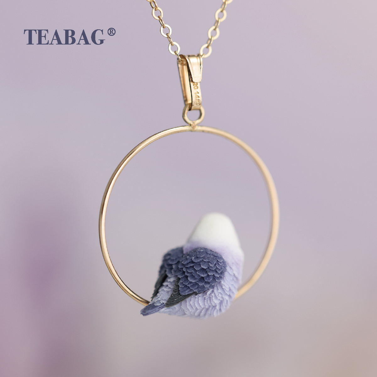 Back view of the Violet Lovebird Necklace with the gold-colored bail and chain connection in focus, above the circular hoop and sculpted purple wings and tail. TEABAG branding is visible.