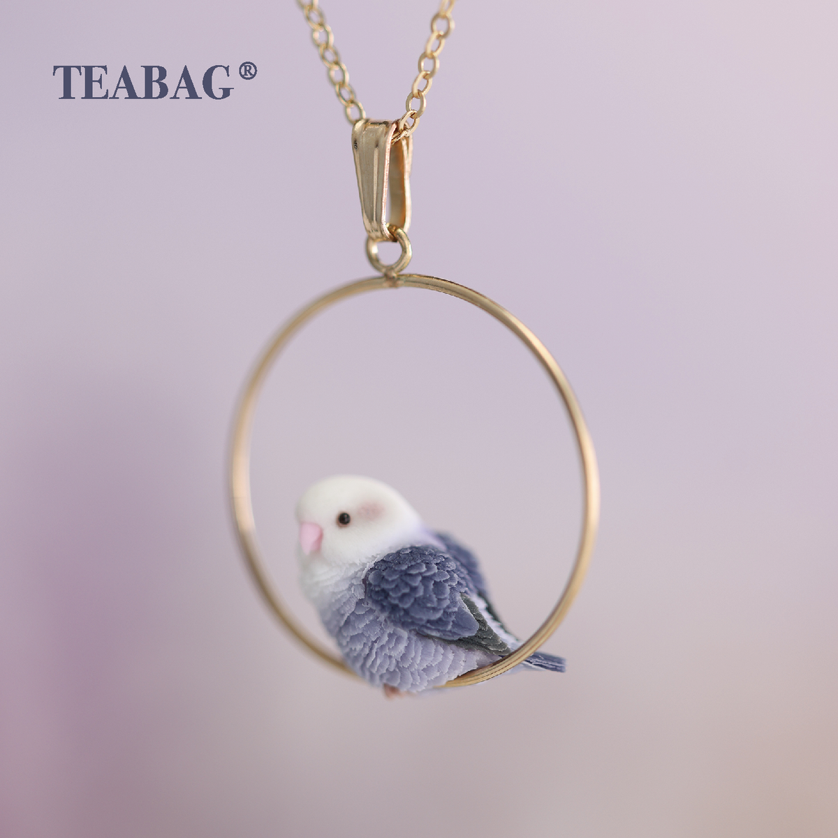 Close view of the Violet Lovebird Necklace with layered purple wing feathers in focus, showing the pale head, gold-colored hoop, bail and chain section against a lavender background.