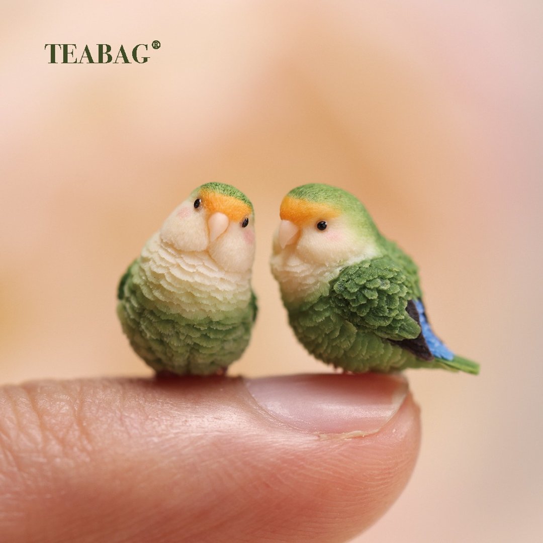 Two miniature green lovebird charms shown together on a fingertip.