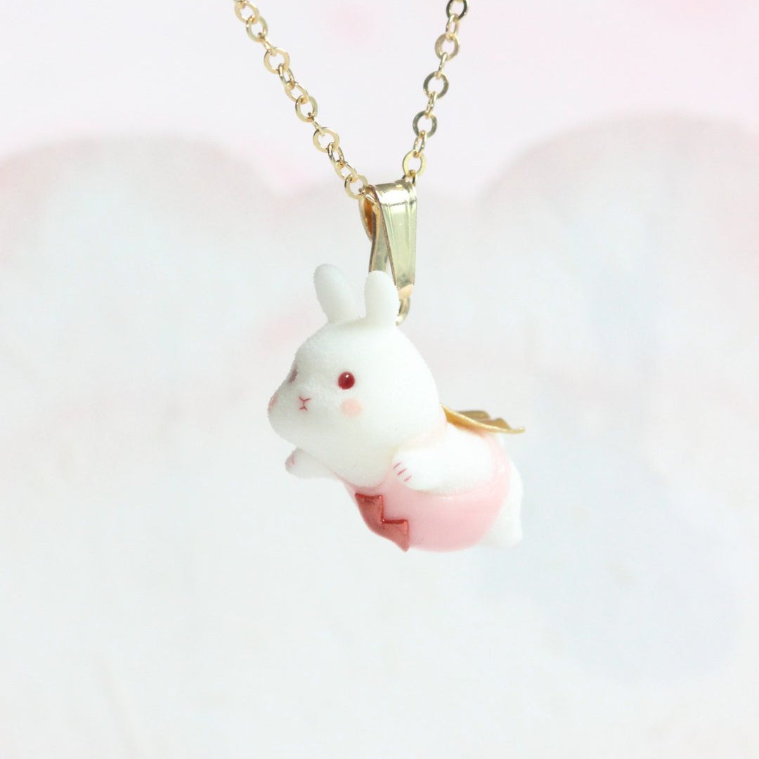 Side view of a white flying-bunny pendant in pink, hanging from a gold-colored chain.