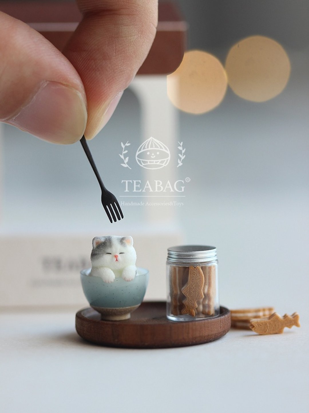 Hand holding a miniature fork above the kitten-and-cookie-jar styling scene.