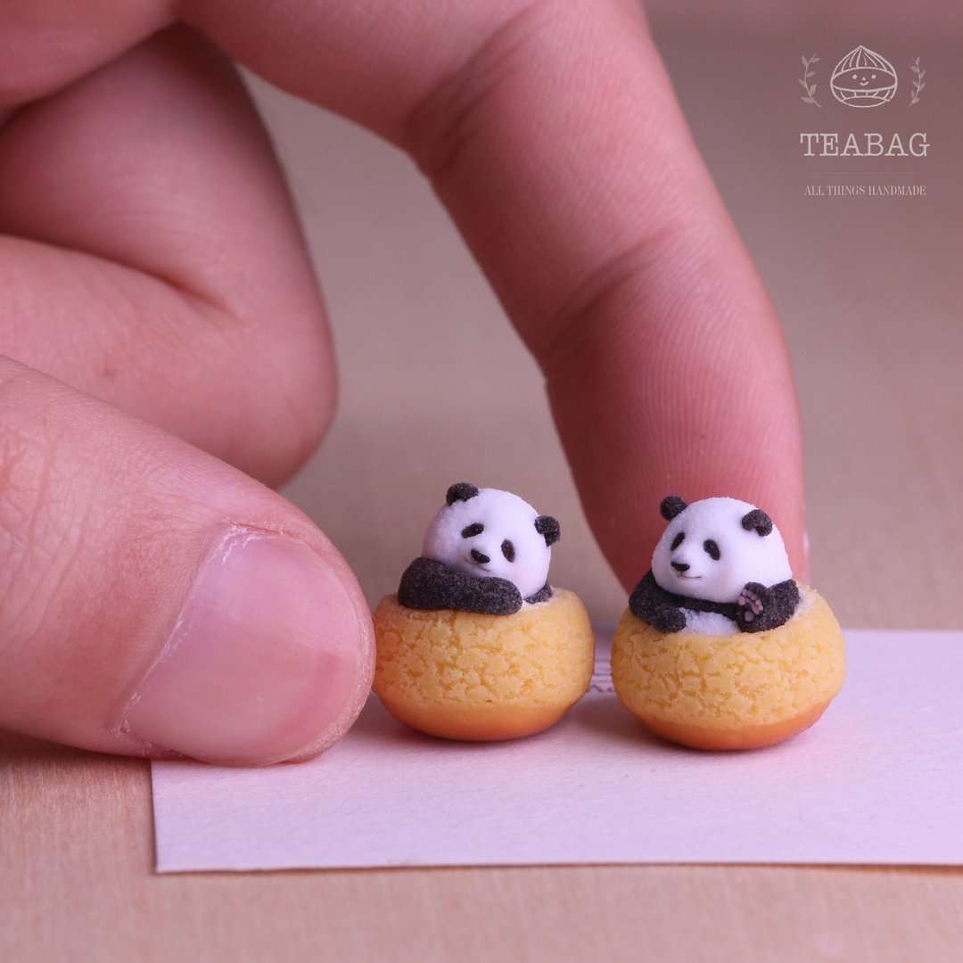 Front view of the Pair: one resting and one waving Panda Cream Puff miniature without jewelry fittings, showing both complete golden pastries, fingertips for scale and the TEABAG logo in a square photograph.