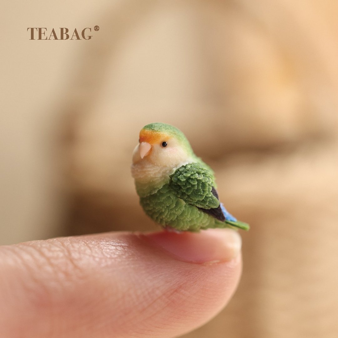 Three-quarter view of the lovebird charm on a fingertip, with a pale face and green wings.