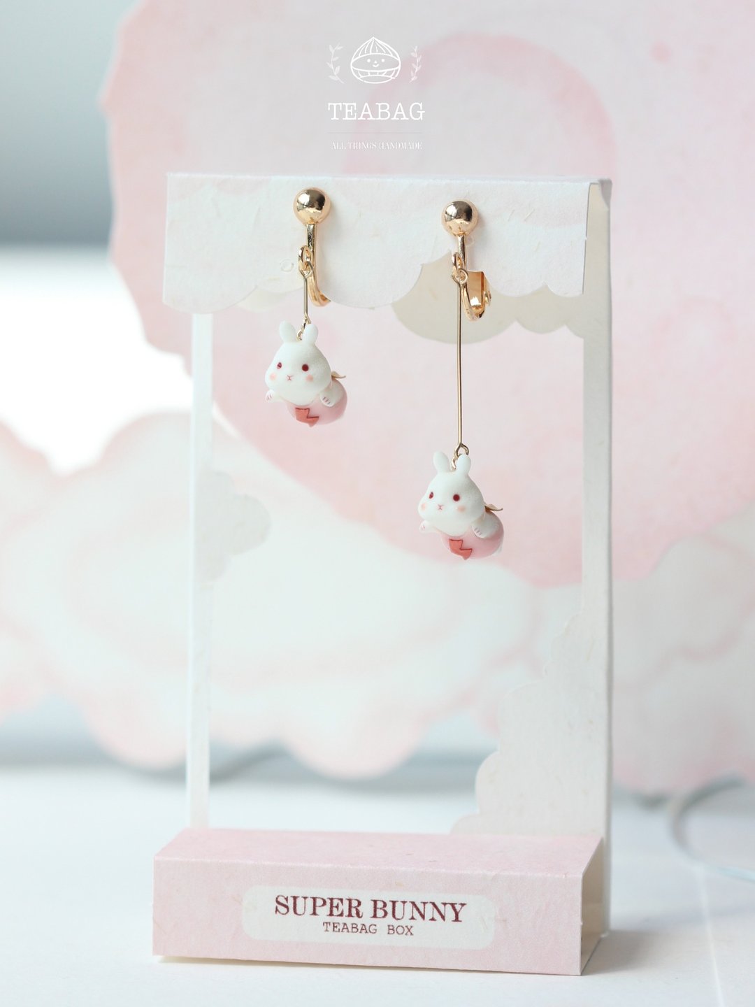 Asymmetric flying-bunny earrings displayed together on a pink card.