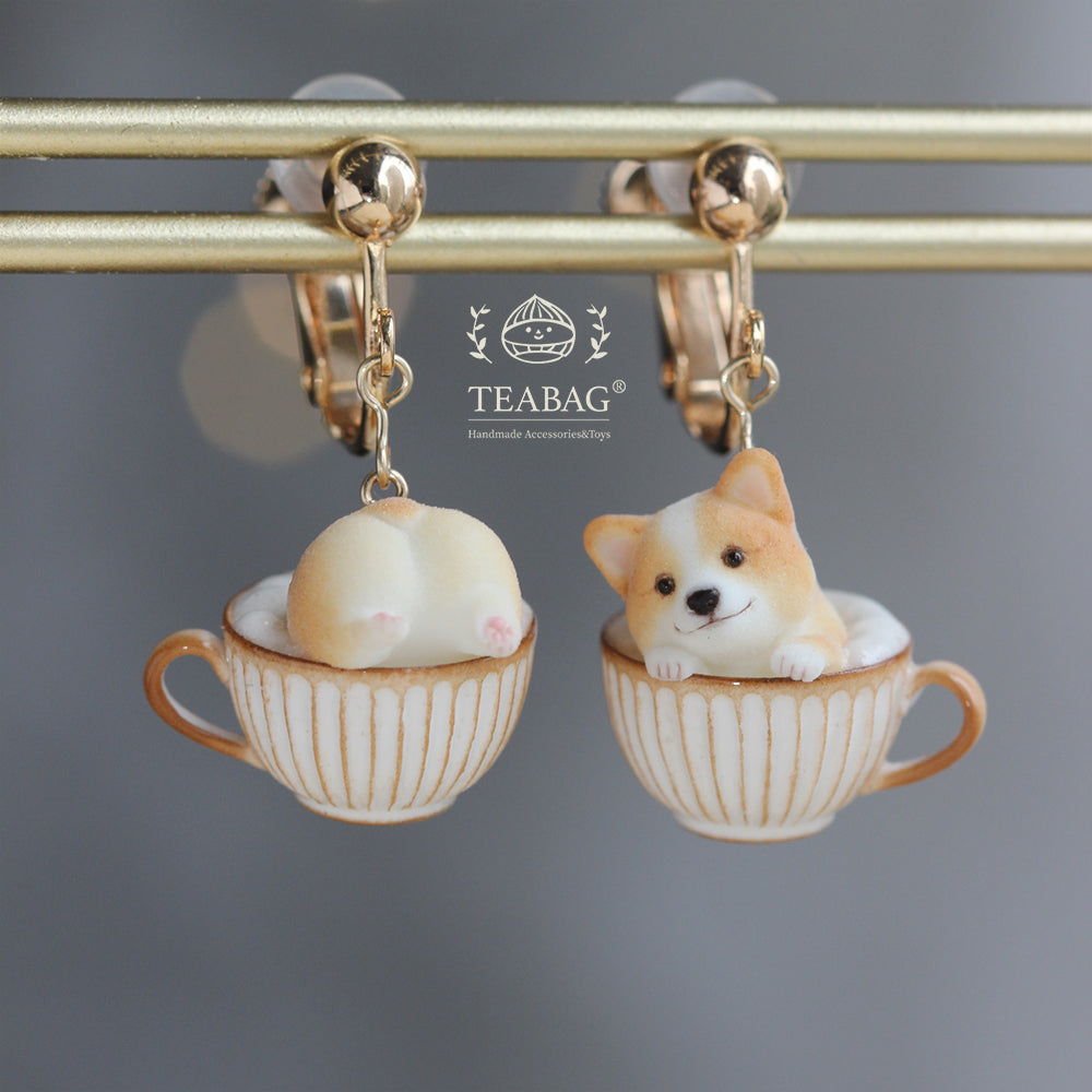 Front product view of the complete Corgi Coffee clip-on earring pair on a display rail, showing both clip fittings, the matching cream-and-gold cups and the TEABAG logo. Not worn on ears.