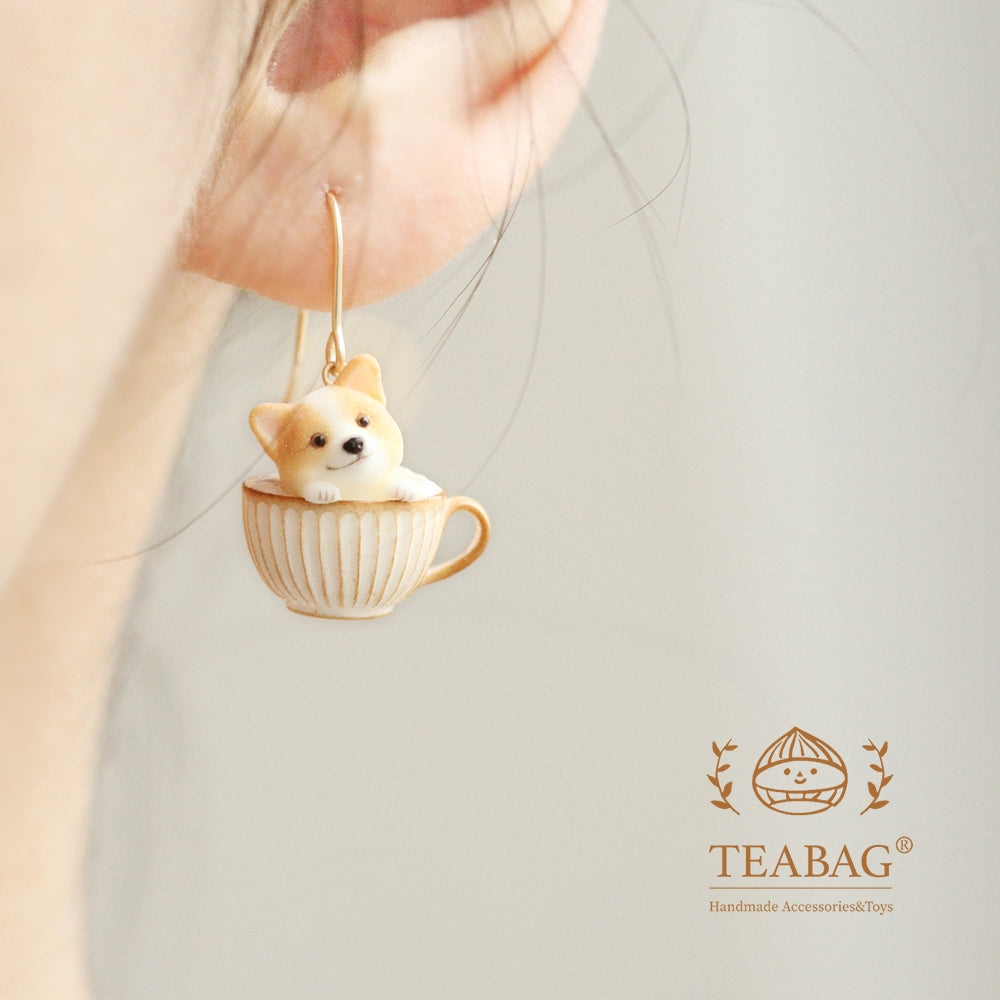 Square photograph of the front-facing Corgi Coffee pierced-hook earring worn on an ear, with the complete hook, cream-and-gold cup and TEABAG logo visible.