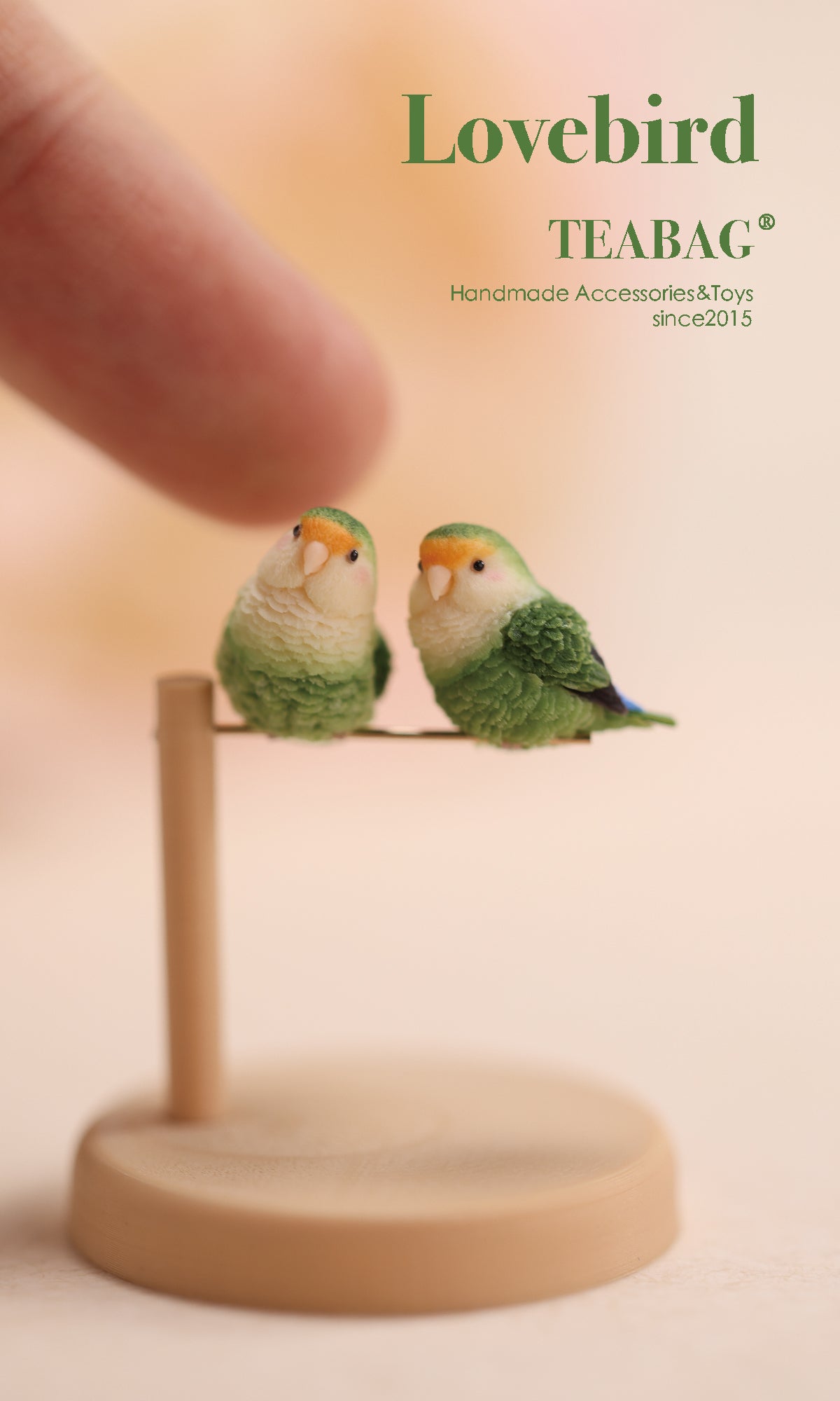 Two handmade miniature green lovebirds on one display stand, with a fingertip above them for scale and TEABAG branding.