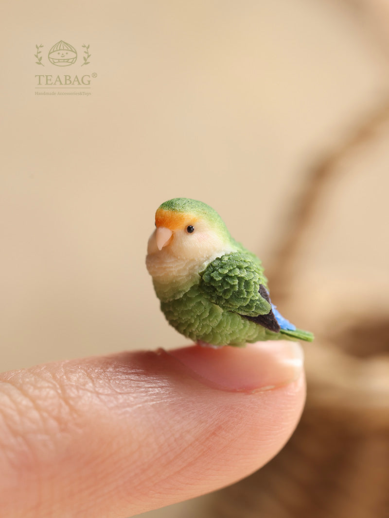 Portrait view of a side-facing green lovebird miniature on a real fingertip for scale, showing sculpted feathers and TEABAG branding.