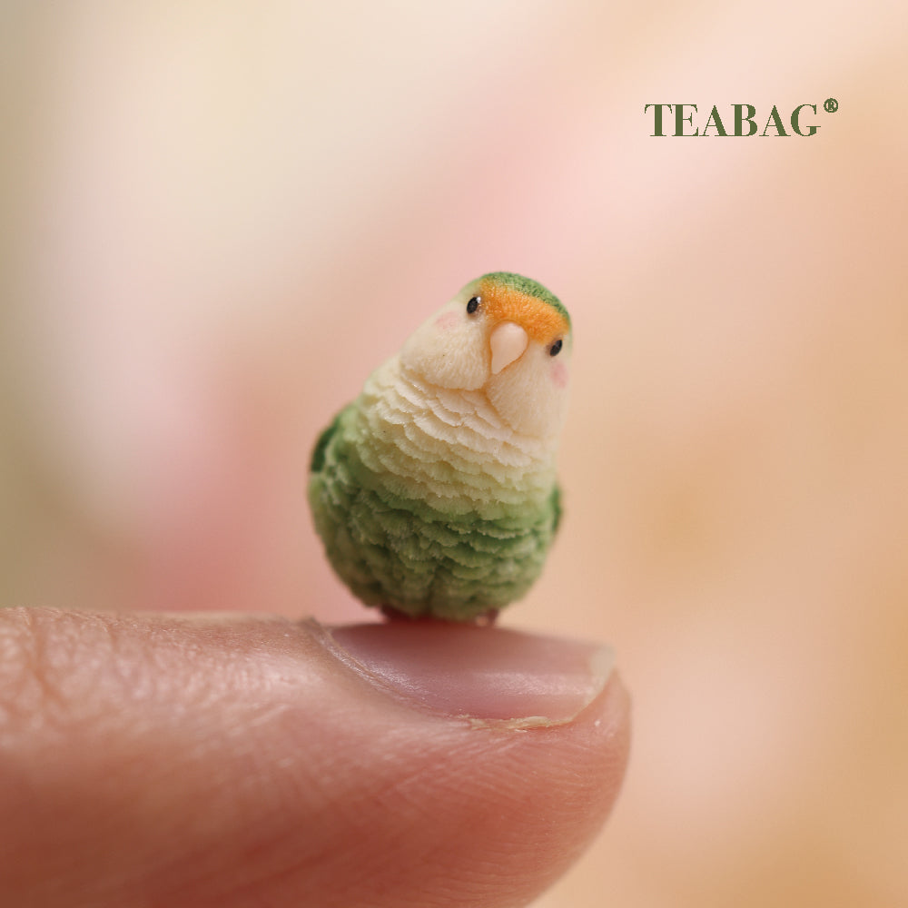 Tilted-head green lovebird miniature resting on a fingertip for scale, with TEABAG branding.